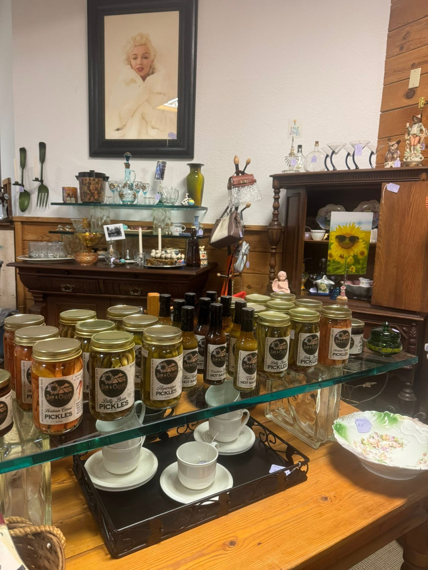 Jars of Sun & Cellar pickles and hot sauces on a glass shelf among the antiques.