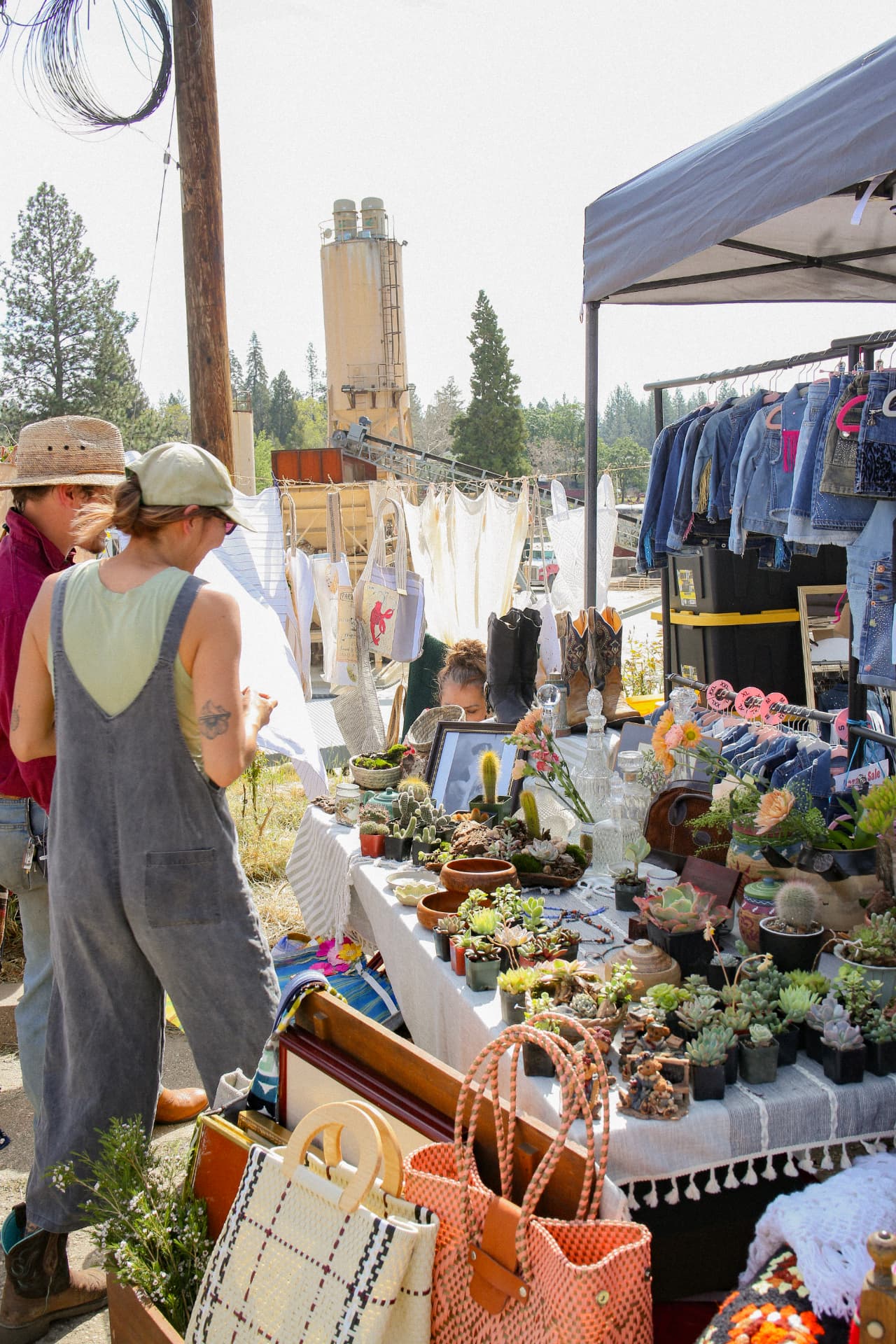 Shoppers browsing a stall of succulents, woven bags, and vintage denim with the old silo behind.